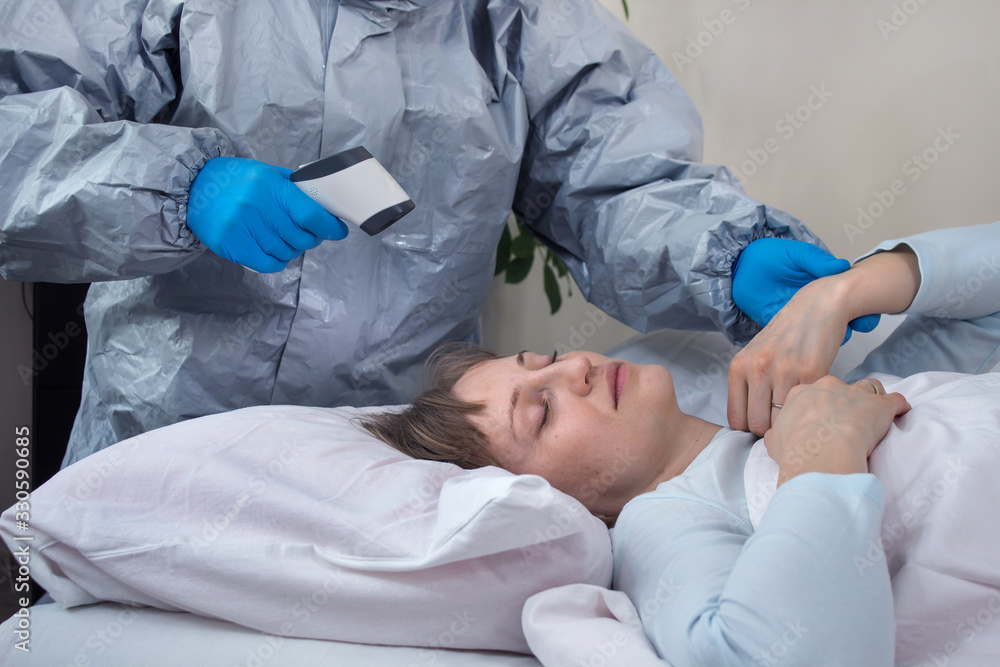 Chora kobieta leży w łóżku. Osoba ubrana w kombinezon ochronny i gumowe rękawiczki mierzy kobiecie temperaturę za pomocą termometru laserowego. Drugą ręką mierzy puls chorej osobie. Pomoc osobom w kwa