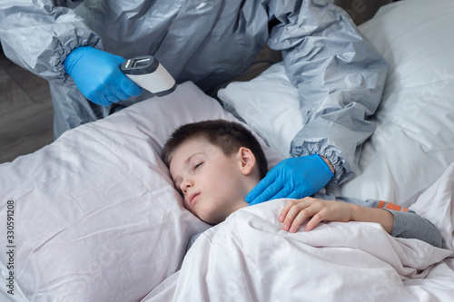 Na pierwszym planie dłoń trzyma termometr elektroniczny gotowy do pomiaru. W tle twarz chłopca śpiącego w łóżku na białej pościeli. 
