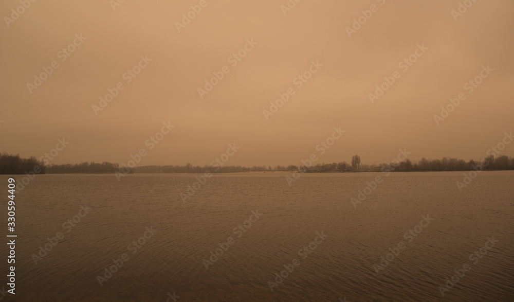 A wide landscape of a dark gray lake and forest. Overcast sky