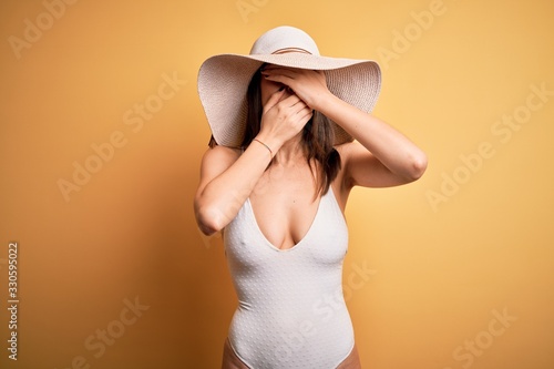 Young beautiful brunette woman on vacation wearing swimsuit and summer hat Covering eyes and mouth with hands, surprised and shocked. Hiding emotion