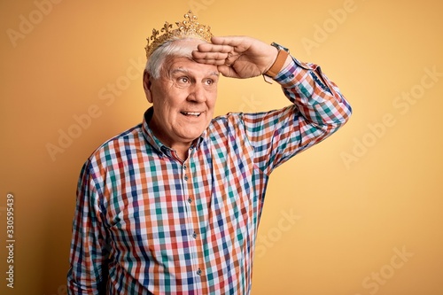 Senior handsome hoary man wearing golden crown of king over isolated yellow background very happy and smiling looking far away with hand over head. Searching concept. © Krakenimages.com