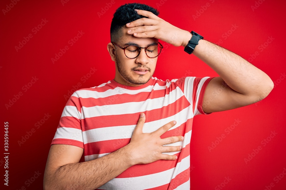 Fototapeta premium Young handsome man wearing casual striped t-shirt and glasses over isolated red background Touching forehead for illness and fever, flu and cold, virus sick