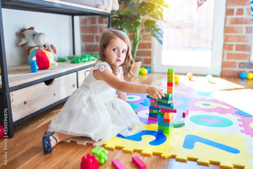 Adorable blonde toddler playing with building blocks toy around lots of toys at kindergarten