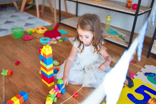 Adorable blonde toddler playing with building blocks around lots of toys at kindergarten
