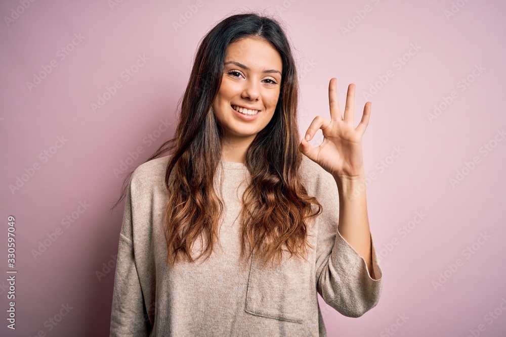 Young beautiful brunette woman wearing casual sweater standing over pink background smiling positive doing ok sign with hand and fingers. Successful expression. Young beautiful brunette woman wearing casual sweater standing over pink background smiling positive doing ok sign with hand and fingers. Successful expression.