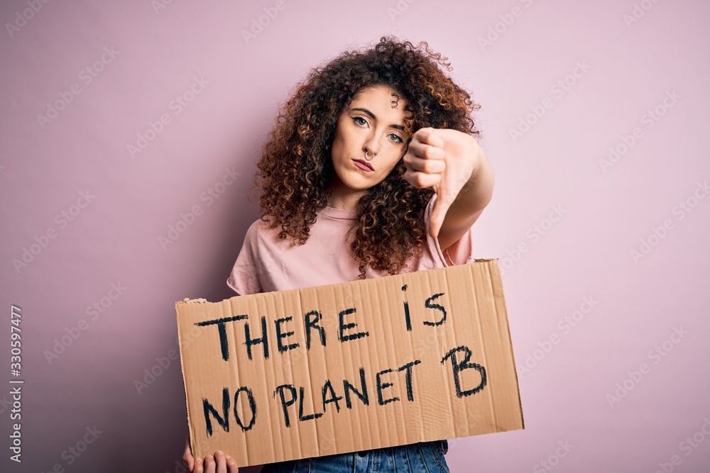 Young beautiful activist woman with curly hair and piercing protesting asking for change planet with angry face, negative sign showing dislike with thumbs down, rejection concept Young beautiful activist woman with curly hair and piercing protesting asking for change planet with angry face, negative sign showing dislike with thumbs down, rejection concept