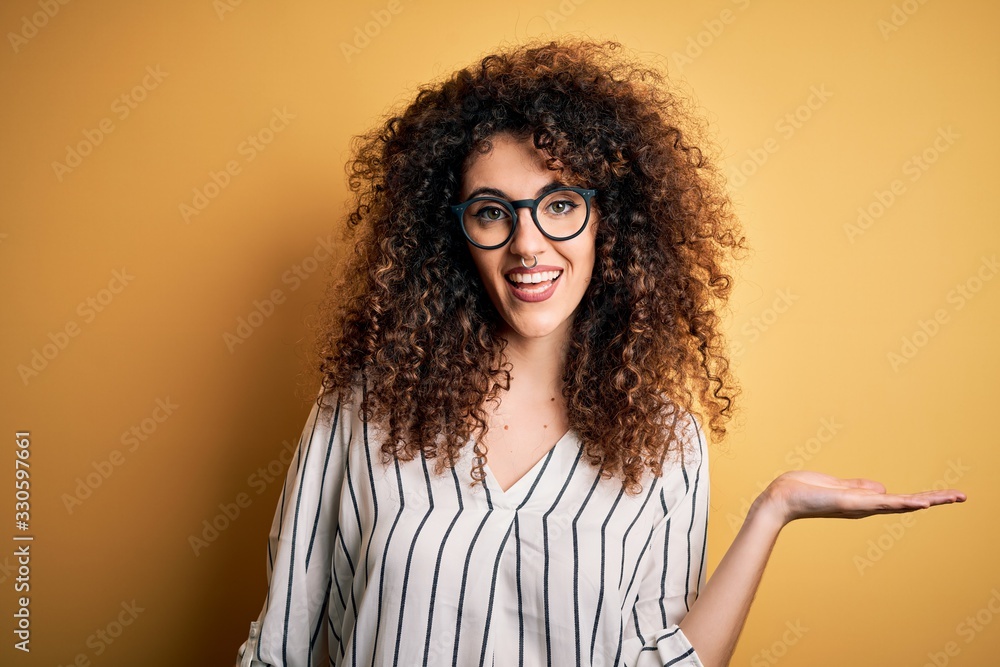 Young beautiful woman with curly hair and piercing wearing striped shirt and glasses smiling cheerful presenting and pointing with palm of hand looking at the camera. Young beautiful woman with curly hair and piercing wearing striped shirt and glasses smiling cheerful presenting and pointing with palm of hand looking at the camera.