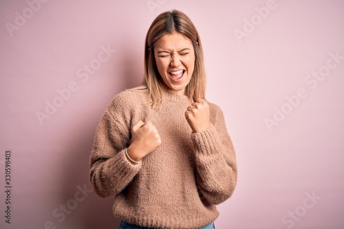 Young beautiful blonde woman wearing winter wool sweater over pink isolated background celebrating surprised and amazed for success with arms raised and eyes closed. Winner concept. © Krakenimages.com