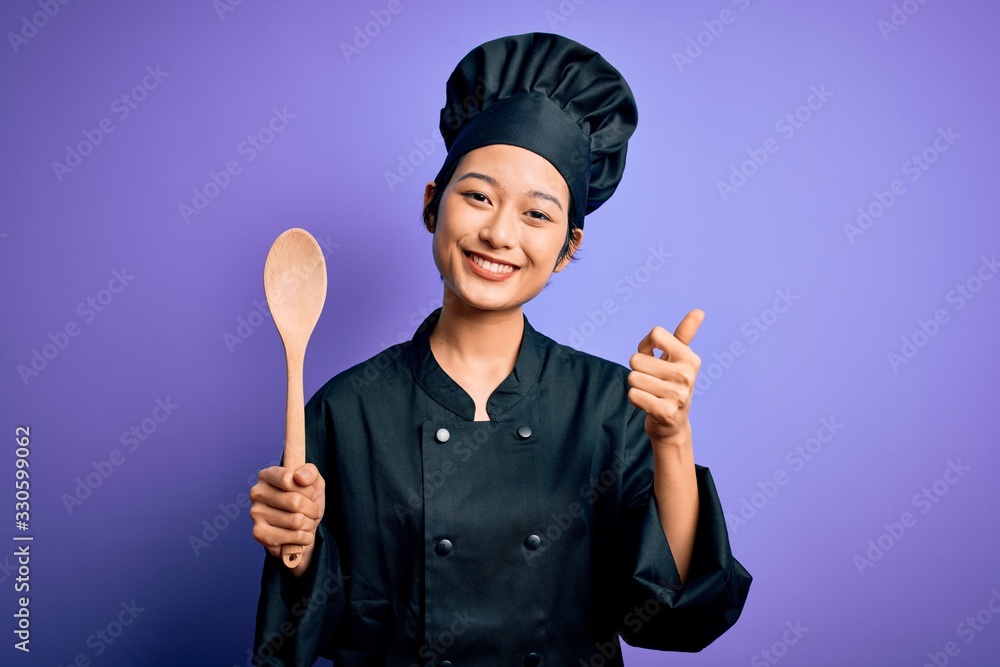 Fototapeta premium Young beautiful chinese chef woman wearing cooker uniform and hat holding wooden spoon happy with big smile doing ok sign, thumb up with fingers, excellent sign