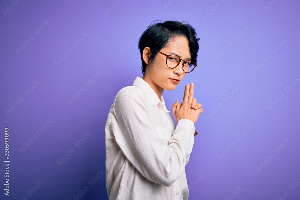Young beautiful asian girl wearing casual shirt and glasses standing over purple background Holding symbolic gun with hand gesture, playing killing shooting weapons, angry face Young beautiful asian girl wearing casual shirt and glasses standing over purple background Holding symbolic gun with hand gesture, playing killing shooting weapons, angry face