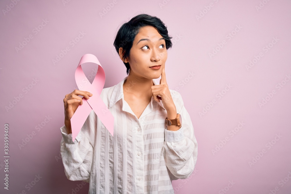 Young beautiful asian girl holding pink cancer ribbon symbol over isolated background serious face thinking about question, very confused idea Young beautiful asian girl holding pink cancer ribbon symbol over isolated background serious face thinking about question, very confused idea