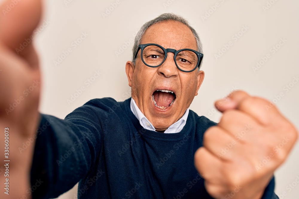 Senior handsome grey-haired man wearing sweater and glasses making selfie by camera annoyed and frustrated shouting with anger, crazy and yelling with raised hand, anger concept Senior handsome grey-haired man wearing sweater and glasses making selfie by camera annoyed and frustrated shouting with anger, crazy and yelling with raised hand, anger concept
