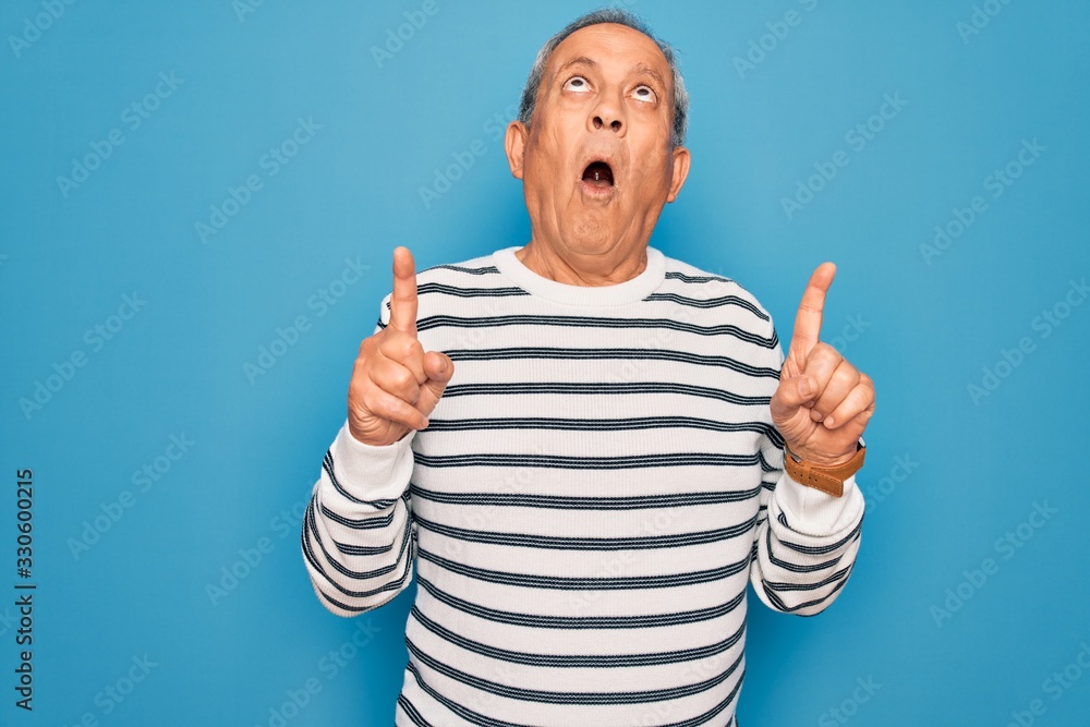 Senior handsome grey-haired man wearing striped sweater over isolated blue background amazed and surprised looking up and pointing with fingers and raised arms. Senior handsome grey-haired man wearing striped sweater over isolated blue background amazed and surprised looking up and pointing with fingers and raised arms.