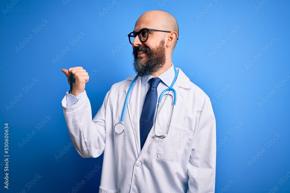 Handsome bald doctor man with beard wearing glasses and stethoscope over blue background smiling with happy face looking and pointing to the side with thumb up. Handsome bald doctor man with beard wearing glasses and stethoscope over blue background smiling with happy face looking and pointing to the side with thumb up.