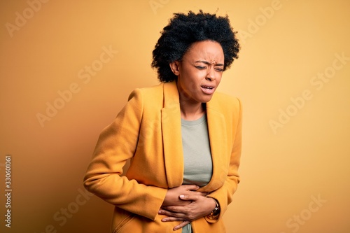Young beautiful African American afro businesswoman with curly hair wearing yellow jacket with hand on stomach because nausea, painful disease feeling unwell. Ache concept. © Krakenimages.com