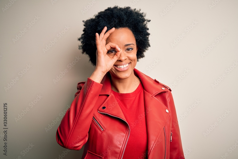 Young beautiful African American afro woman with curly hair wearing casual red jacket doing ok gesture with hand smiling, eye looking through fingers with happy face. Young beautiful African American afro woman with curly hair wearing casual red jacket doing ok gesture with hand smiling, eye looking through fingers with happy face.