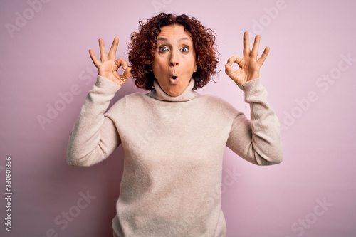 Middle age beautiful curly hair woman wearing casual turtleneck sweater over pink background looking surprised and shocked doing ok approval symbol with fingers. Crazy expression