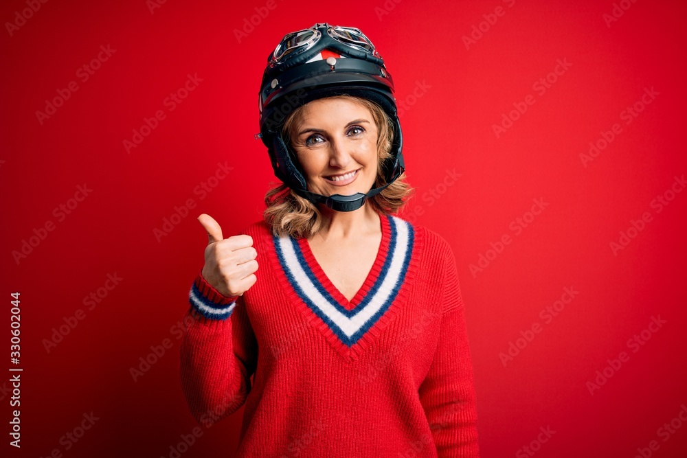 Middle age beautiful blonde motorcyclist woman wearing moto helmet over red background doing happy thumbs up gesture with hand. Approving expression looking at the camera showing success. Middle age beautiful blonde motorcyclist woman wearing moto helmet over red background doing happy thumbs up gesture with hand. Approving expression looking at the camera showing success.