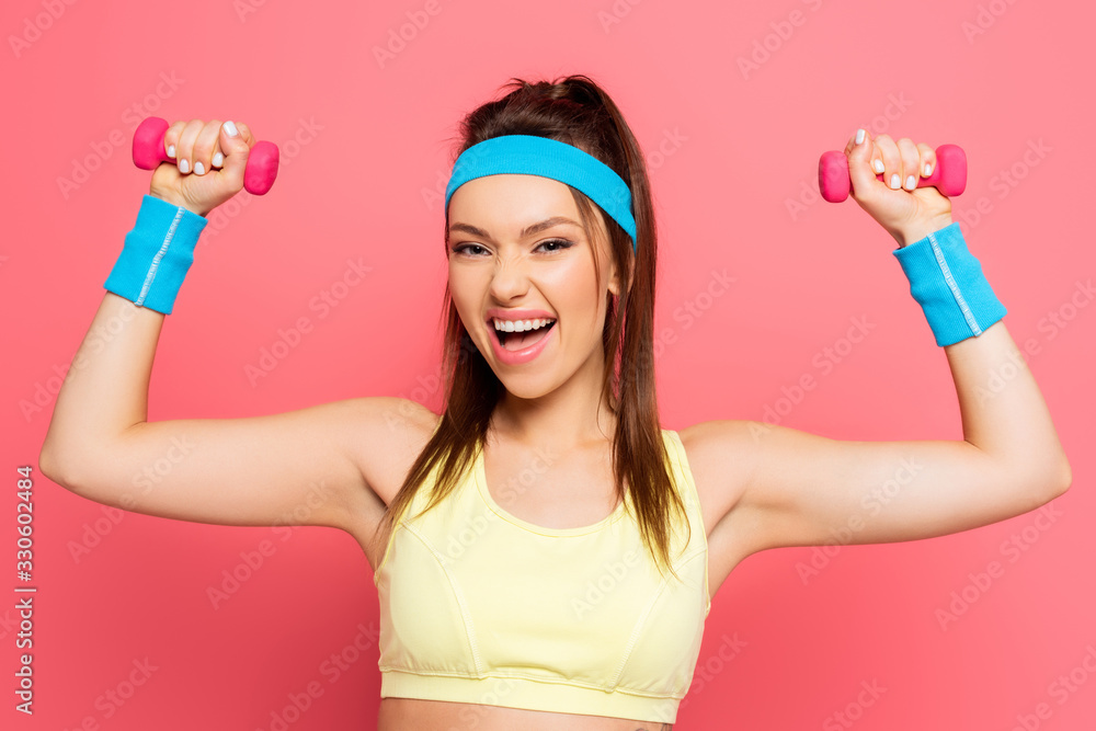 excited sportswoman training with dumbbells while looking at camera on pink background excited sportswoman training with dumbbells while looking at camera on pink background