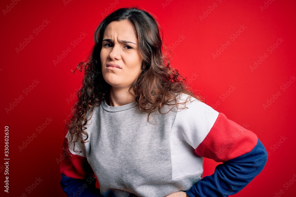Young beautiful woman with curly hair wearing casual sweatshirt over isolated red background with hand on stomach because indigestion, painful illness feeling unwell. Ache concept. Young beautiful woman with curly hair wearing casual sweatshirt over isolated red background with hand on stomach because indigestion, painful illness feeling unwell. Ache concept.