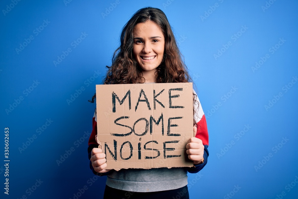 Beautiful activist woman holding banner with make some noise message over blue background with a happy face standing and smiling with a confident smile showing teeth Beautiful activist woman holding banner with make some noise message over blue background with a happy face standing and smiling with a confident smile showing teeth