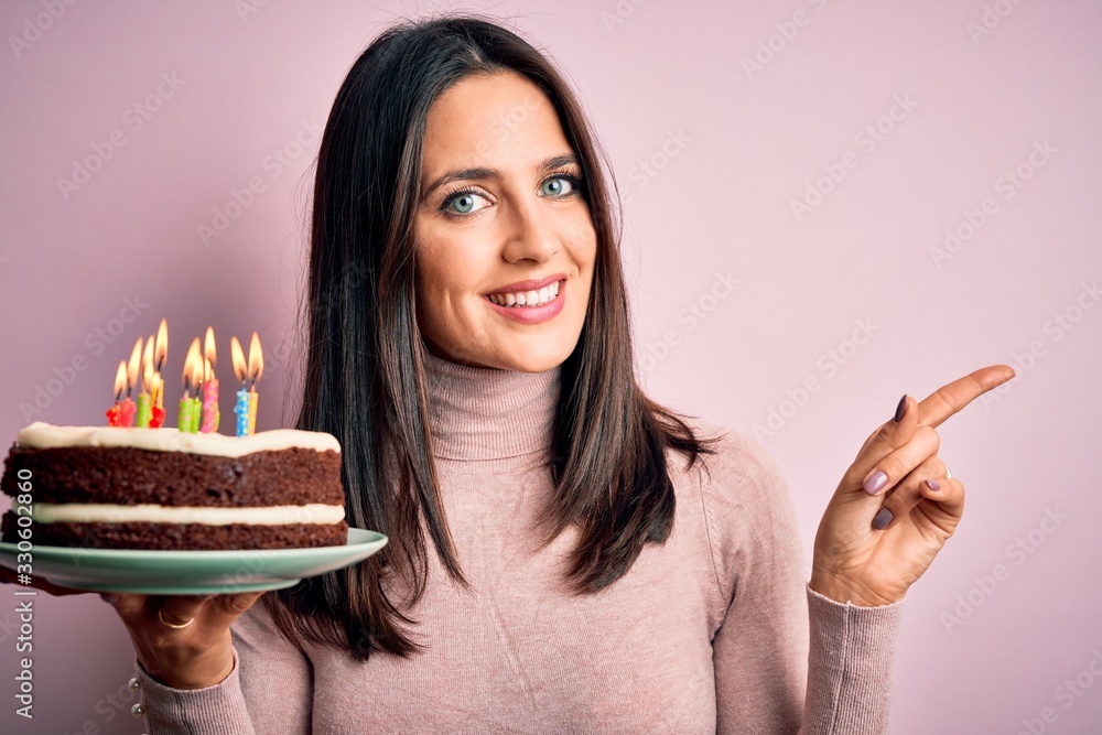 Young woman with blue eyes holding birthday cake with candles over pink background very happy pointing with hand and finger to the side Young woman with blue eyes holding birthday cake with candles over pink background very happy pointing with hand and finger to the side