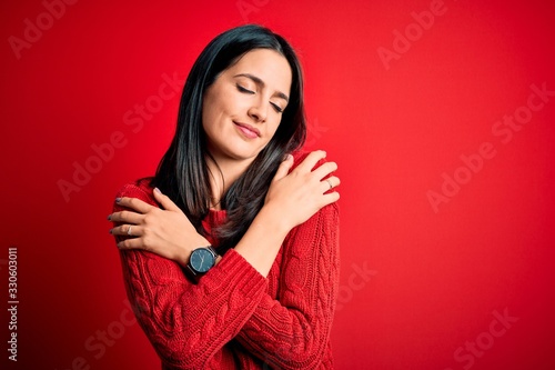 Young brunette woman with blue eyes wearing casual sweater over isolated red background Hugging oneself happy and positive, smiling confident. Self love and self care