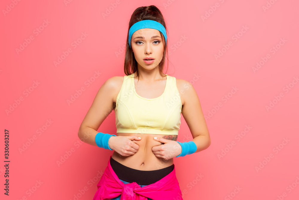 displeased sportswoman touching her body and looking at camera on pink background displeased sportswoman touching her body and looking at camera on pink background