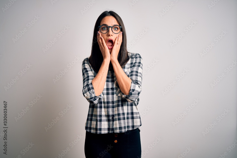 Young brunette woman with blue eyes wearing casual shirt and glasses over white background afraid and shocked, surprise and amazed expression with hands on face Young brunette woman with blue eyes wearing casual shirt and glasses over white background afraid and shocked, surprise and amazed expression with hands on face