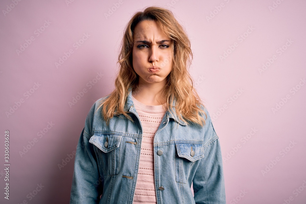 Young beautiful blonde woman wearing casual denim jacket standing over pink background puffing cheeks with funny face. Mouth inflated with air, crazy expression. Young beautiful blonde woman wearing casual denim jacket standing over pink background puffing cheeks with funny face. Mouth inflated with air, crazy expression.
