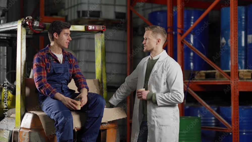 Tracking medium shot of warehouse manager in white coat and forklift driver talking over coffee at break in industrial storage Tracking medium shot of warehouse manager in white coat and forklift driver talking over coffee at break in industrial storage