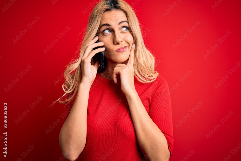 Beautiful blonde woman having conversation talking on the smartphone over red background serious face thinking about question, very confused idea Beautiful blonde woman having conversation talking on the smartphone over red background serious face thinking about question, very confused idea