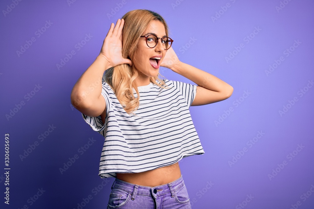 Young beautiful blonde woman wearing striped t-shirt and glasses over purple background Smiling cheerful playing peek a boo with hands showing face. Surprised and exited Young beautiful blonde woman wearing striped t-shirt and glasses over purple background Smiling cheerful playing peek a boo with hands showing face. Surprised and exited