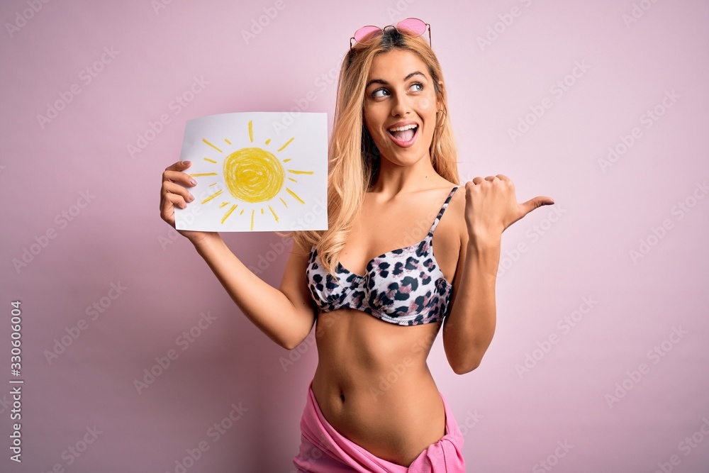 Young beautiful blonde woman on vacation wearing bikini holding banner with sun draw pointing and showing with thumb up to the side with happy face smiling Young beautiful blonde woman on vacation wearing bikini holding banner with sun draw pointing and showing with thumb up to the side with happy face smiling
