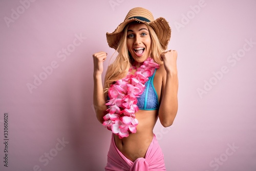 Young beautiful blonde woman on vacation wearing bikini and hat with hawaiian lei flowers celebrating surprised and amazed for success with arms raised and open eyes. Winner concept.