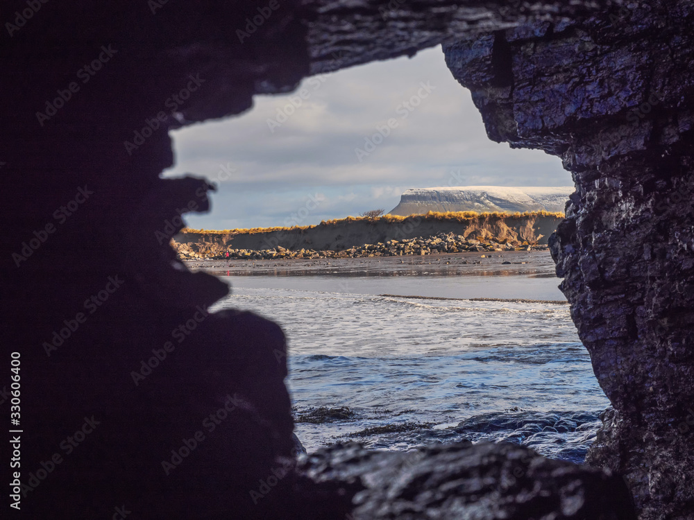 View on Atlantic ocean from a cave by Rosses point beach in county Sligo Ireland, Benbulben mountain covered with snow on the right.