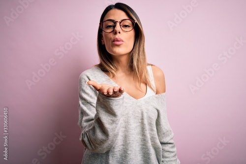 Young beautiful brunette woman wearing casual sweater and glasses over pink background looking at the camera blowing a kiss with hand on air being lovely and sexy. Love expression.