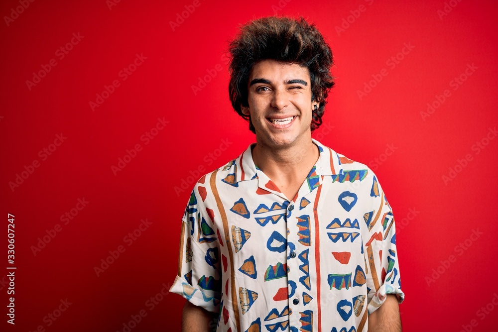 Young handsome man on vacation wearing summer shirt over isolated red background winking looking at the camera with sexy expression, cheerful and happy face. Young handsome man on vacation wearing summer shirt over isolated red background winking looking at the camera with sexy expression, cheerful and happy face.