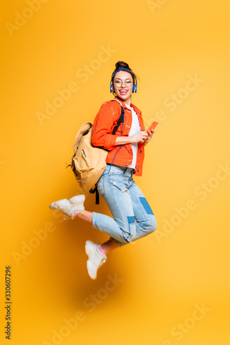 excited student in headset and glasses jumping while holding smartphone on yellow background