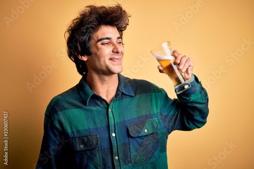 Young handsome man smiling happy. Standing with smile on face drinking glass of beer over isolated yellow background