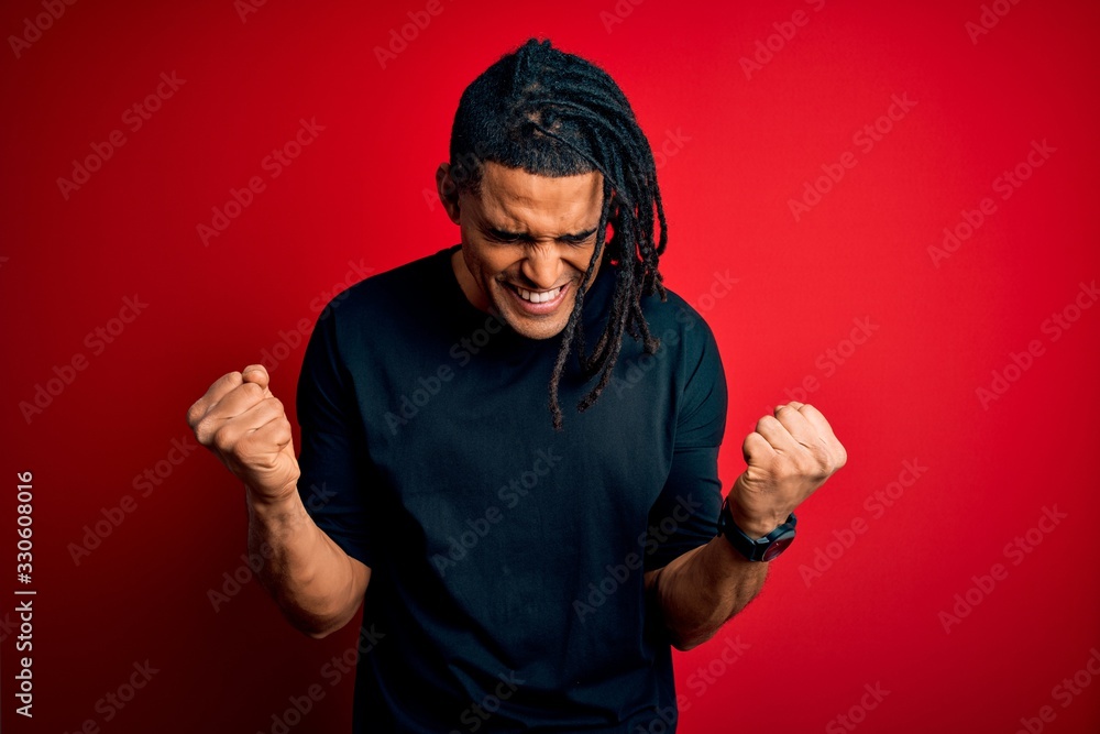 Fototapeta premium Young handsome african american afro man with dreadlocks wearing casual t-shirt very happy and excited doing winner gesture with arms raised, smiling and screaming for success. Celebration concept.