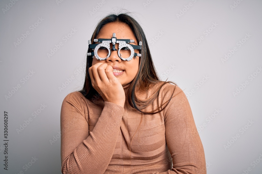 Young asian optical girl controlling eyesight using optometry glasses over white background looking stressed and nervous with hands on mouth biting nails. Anxiety problem. Young asian optical girl controlling eyesight using optometry glasses over white background looking stressed and nervous with hands on mouth biting nails. Anxiety problem.