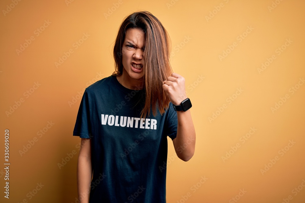 Young beautiful brunette girl doing volunteering wearing t-shirt with volunteer message word angry and mad raising fist frustrated and furious while shouting with anger. Rage and aggressive concept. Young beautiful brunette girl doing volunteering wearing t-shirt with volunteer message word angry and mad raising fist frustrated and furious while shouting with anger. Rage and aggressive concept.