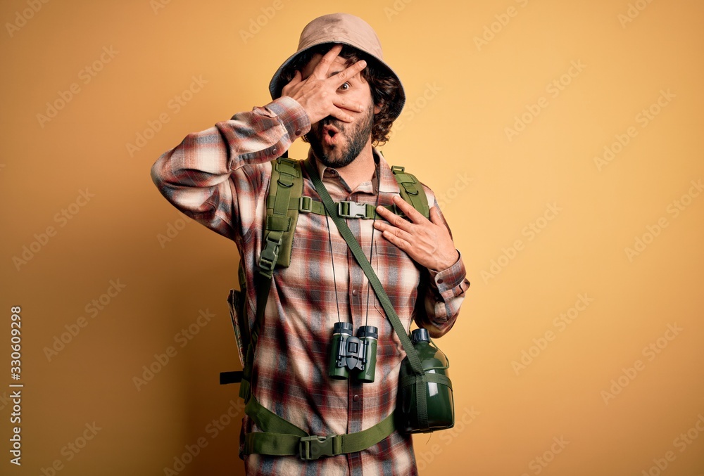 Young hiker man with curly hair and beard hiking wearing backpack and water canteen peeking in shock covering face and eyes with hand, looking through fingers with embarrassed expression. Young hiker man with curly hair and beard hiking wearing backpack and water canteen peeking in shock covering face and eyes with hand, looking through fingers with embarrassed expression.