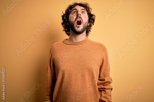 Young handsome man with beard wearing casual sweater standing over yellow background angry and mad screaming frustrated and furious, shouting with anger. Rage and aggressive concept. © Krakenimages.com