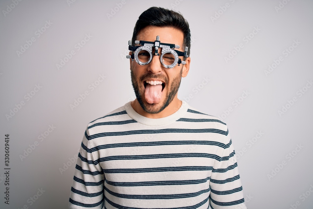 Obraz premium Young handsome man with beard wearing optometry glasses over isolated white background sticking tongue out happy with funny expression. Emotion concept.