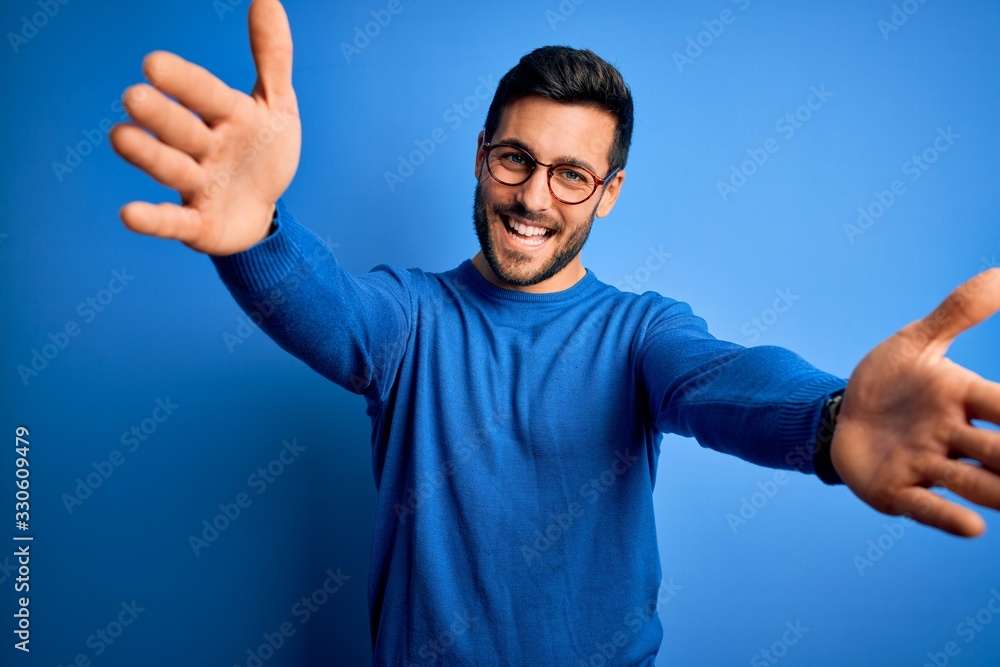 Young handsome man with beard wearing casual sweater and glasses over blue background looking at the camera smiling with open arms for hug. Cheerful expression embracing happiness. Young handsome man with beard wearing casual sweater and glasses over blue background looking at the camera smiling with open arms for hug. Cheerful expression embracing happiness.