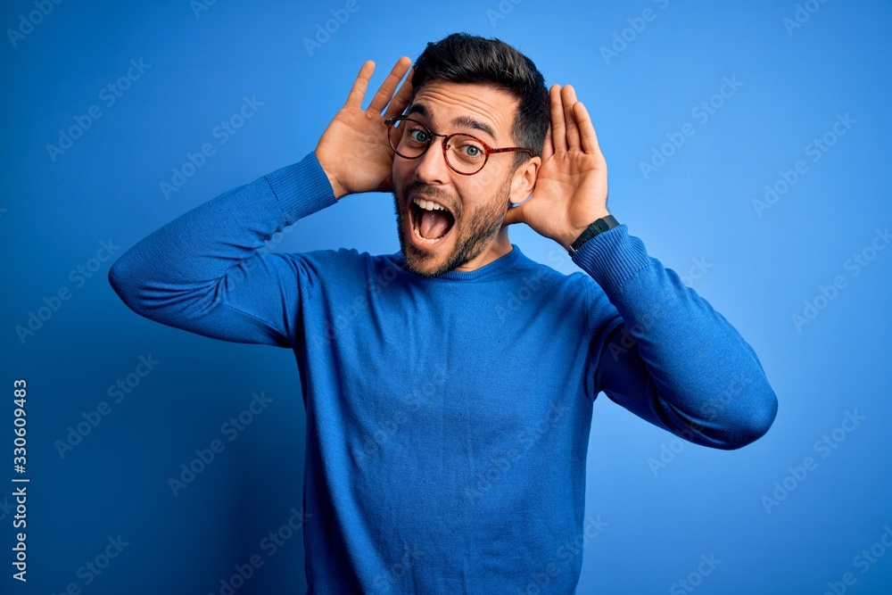 Young handsome man with beard wearing casual sweater and glasses over blue background Smiling cheerful playing peek a boo with hands showing face. Surprised and exited Young handsome man with beard wearing casual sweater and glasses over blue background Smiling cheerful playing peek a boo with hands showing face. Surprised and exited
