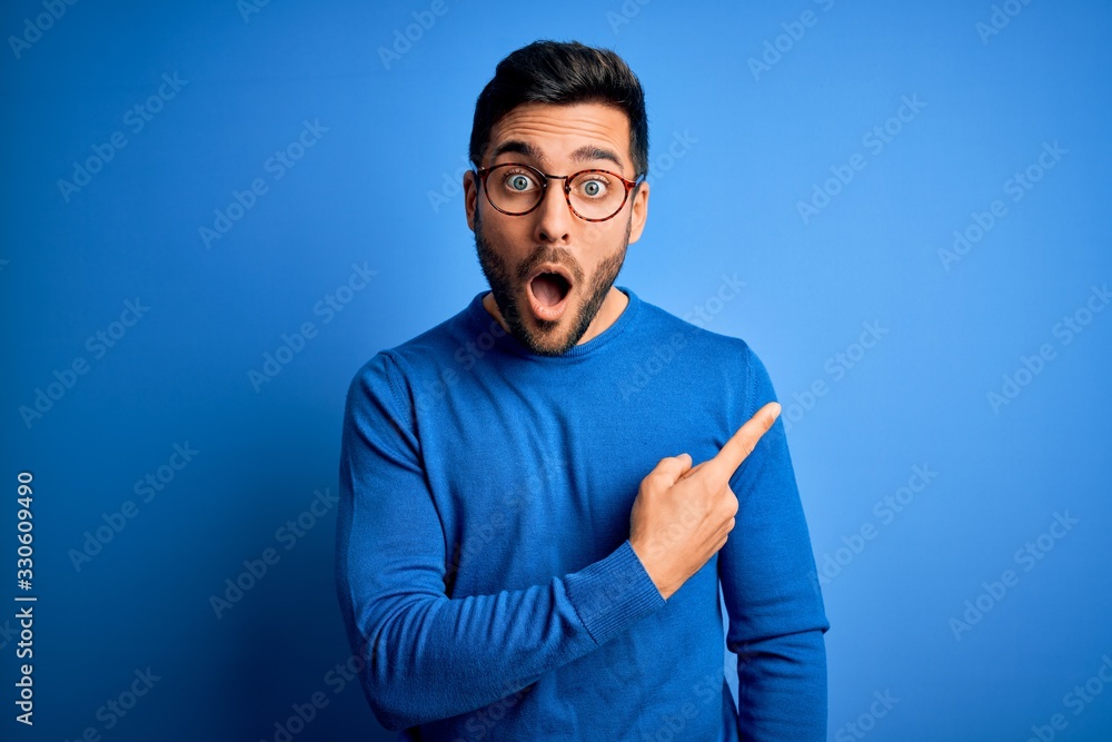 Young handsome man with beard wearing casual sweater and glasses over blue background Surprised pointing with finger to the side, open mouth amazed expression. - obrazy, fototapety, plakaty 