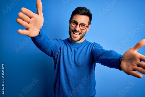 Cuadro en lienzo Young handsome man with beard wearing casual sweater and glasses over blue background looking at the camera smiling with open arms for hug